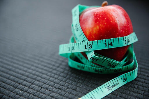 A red apple wrapped in a teal tape measure on a dark textured background, symbolizing weight tracking and body measurement.